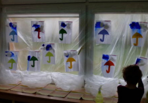 Zafoliowane przedszkolne okno z dziecięcymi pracami plastycznymi przedstawiającymi kolorowy deszcz.